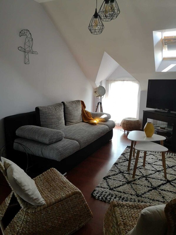 A modern living room with a grey sofa, a small white table, and a TV. There are wicker chairs and a patterned rug.