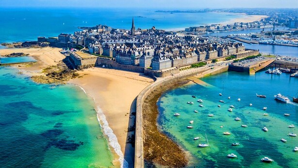 An aerial view of a coastal city with a prominent castle, a sandy beach, and a marina with numerous boats.