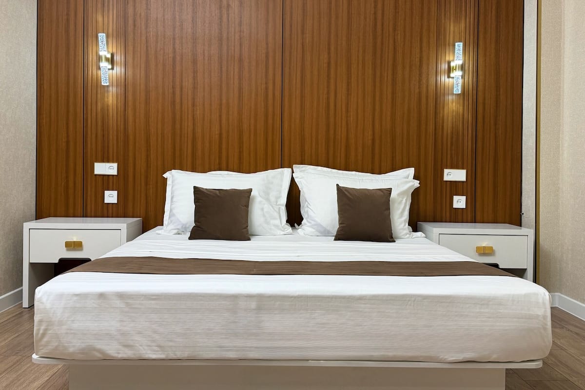 A neatly made bed with white and brown bedding, flanked by two bedside tables with drawers, in a room with wooden paneling on the walls.