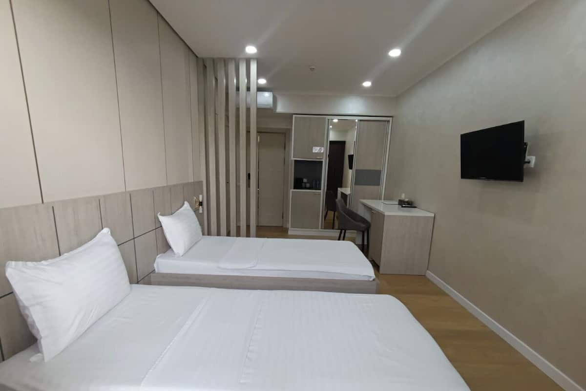 A modern hotel room with two single beds, a flat-screen TV mounted on the wall, and a small desk with a chair.