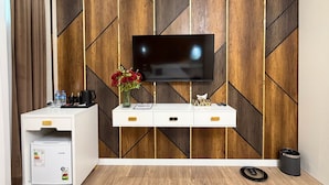 Une chambre d’hôtel moderne dotée d’un téléviseur à écran plat fixé à un mur en bois orné de motifs géométriques, d’un mini-bar blanc, d’un petit réfrigérateur et d’un panier pour animaux placé sur le sol.
