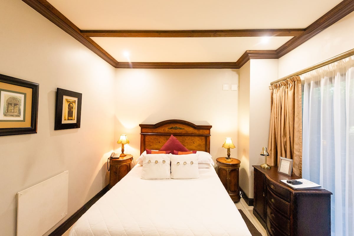 A hotel room with a large bed, wooden headboard, two bedside tables with lamps, a nightstand, a dresser, and a window with curtains.