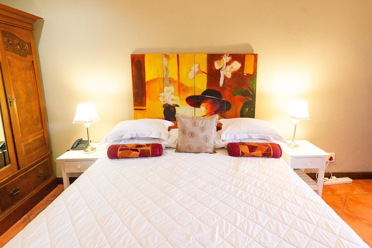 A neatly made bed with a quilted white duvet, two white pillows, and a decorative pillow with a geometric pattern. There are two bedside lamps on either side of the bed, and a wooden wardrobe is visible to the left.