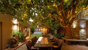 Un patio al aire libre con una mesa y sillas para comer, rodeado de follaje e iluminado con luces colgantes.