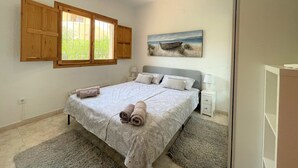 A bedroom with a bed, a window with wooden shutters, a painting of a beach scene, and a small bedside table with a lamp. - T-0819, Villa Sueños, beautiful villa with mediterranean garden (Alicante)
