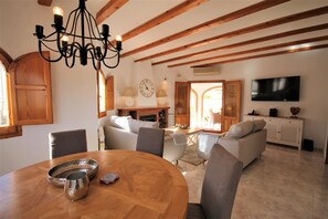 A dining area with a wooden table, chairs, and a chandelier. There is a living room with sofas and a television in the background. - T-0819, Villa Sueños, beautiful villa with mediterranean garden (Alicante)