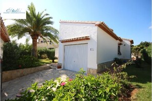 A white building with a tiled roof, a closed white door, and a small garden with a palm tree and flowering plants. - T-0819, Villa Sueños, beautiful villa with mediterranean garden (Alicante)