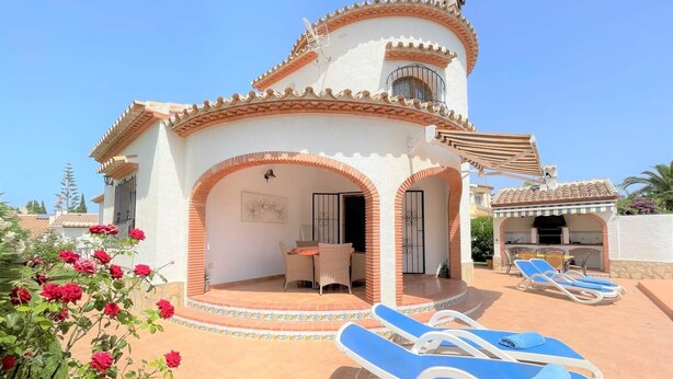 A white villa with a tiled roof, a patio with lounge chairs, and a garden with red flowers. - T-0819, Villa Sueños, beautiful villa with mediterranean garden (Alicante)