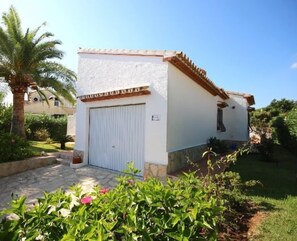 A white building with a tiled roof, a closed white door, and a small garden with a palm tree and flowering plants. - T-0819, Villa Sueños, beautiful villa with mediterranean garden (Alicante)