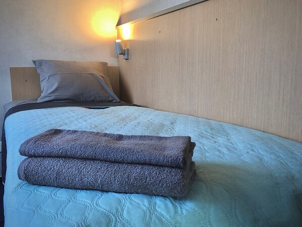 Una cama bien tendida con un almohadón, una colcha doblada y una lámpara fijada a la pared.