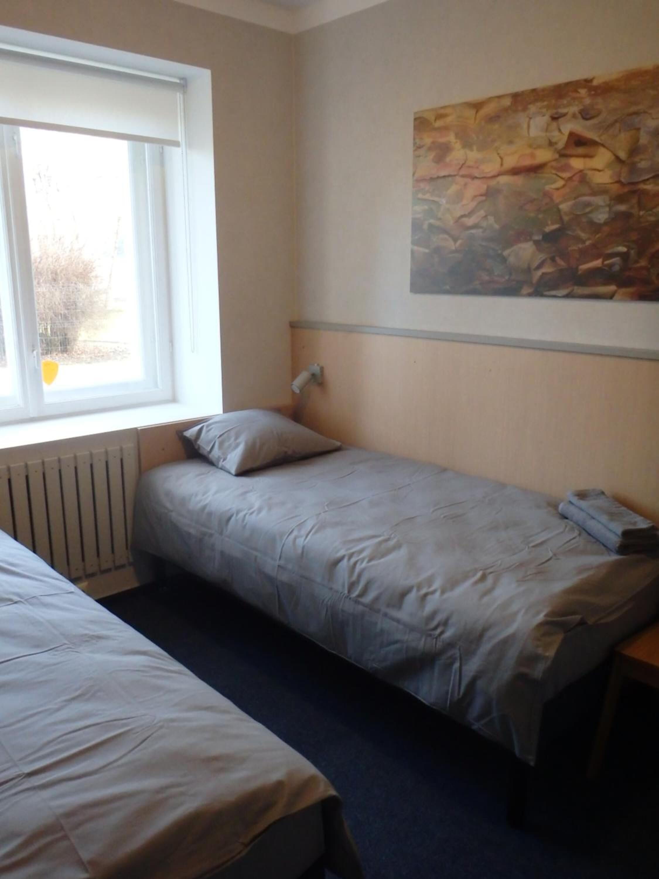 Un petit logement composé d’une seule pièce, comprenant deux lits, un radiateur, une fenêtre et un tableau au mur.