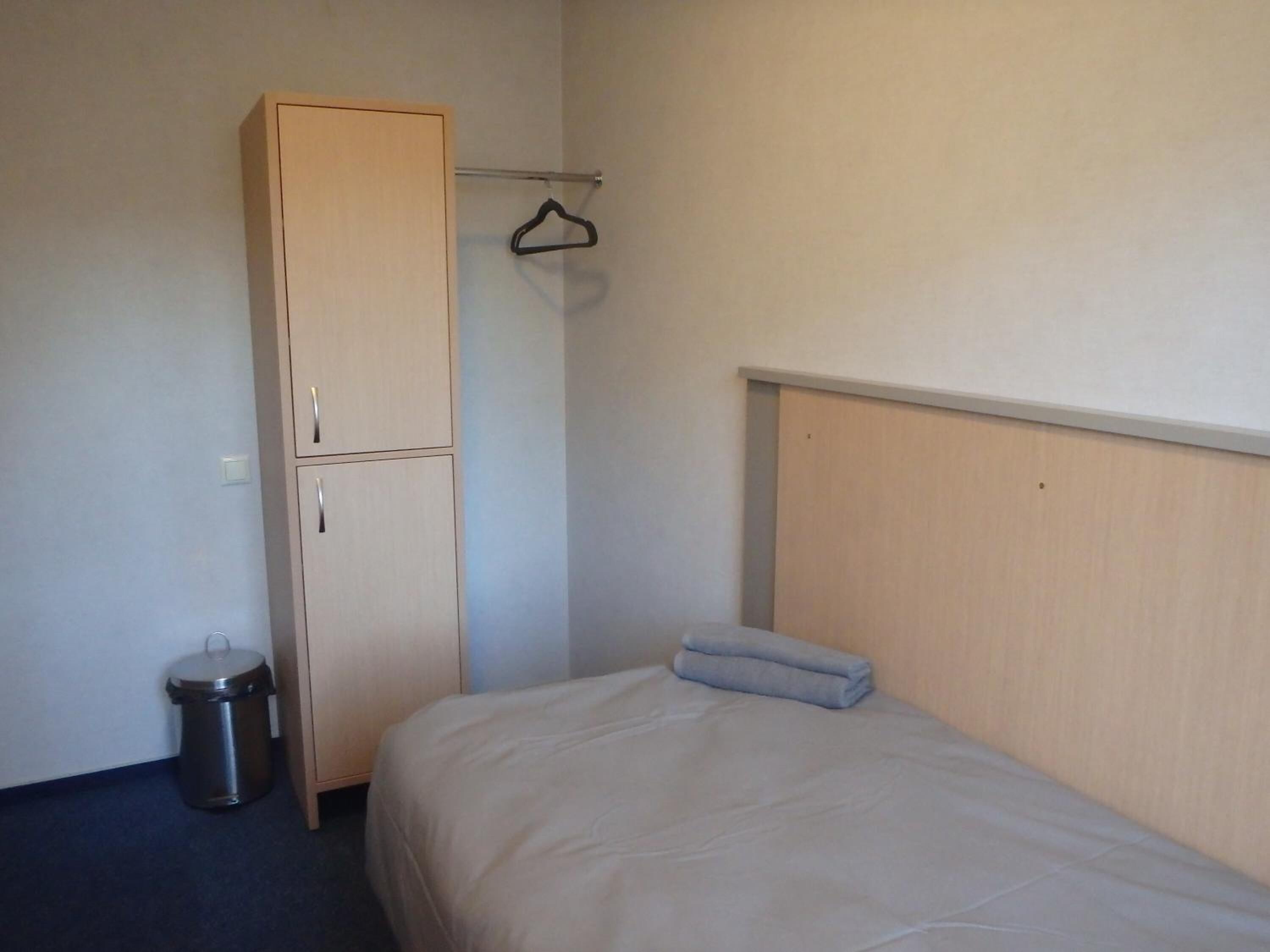 A small, single-bed room with a wooden wardrobe, a hanging rack, and a trash bin.
