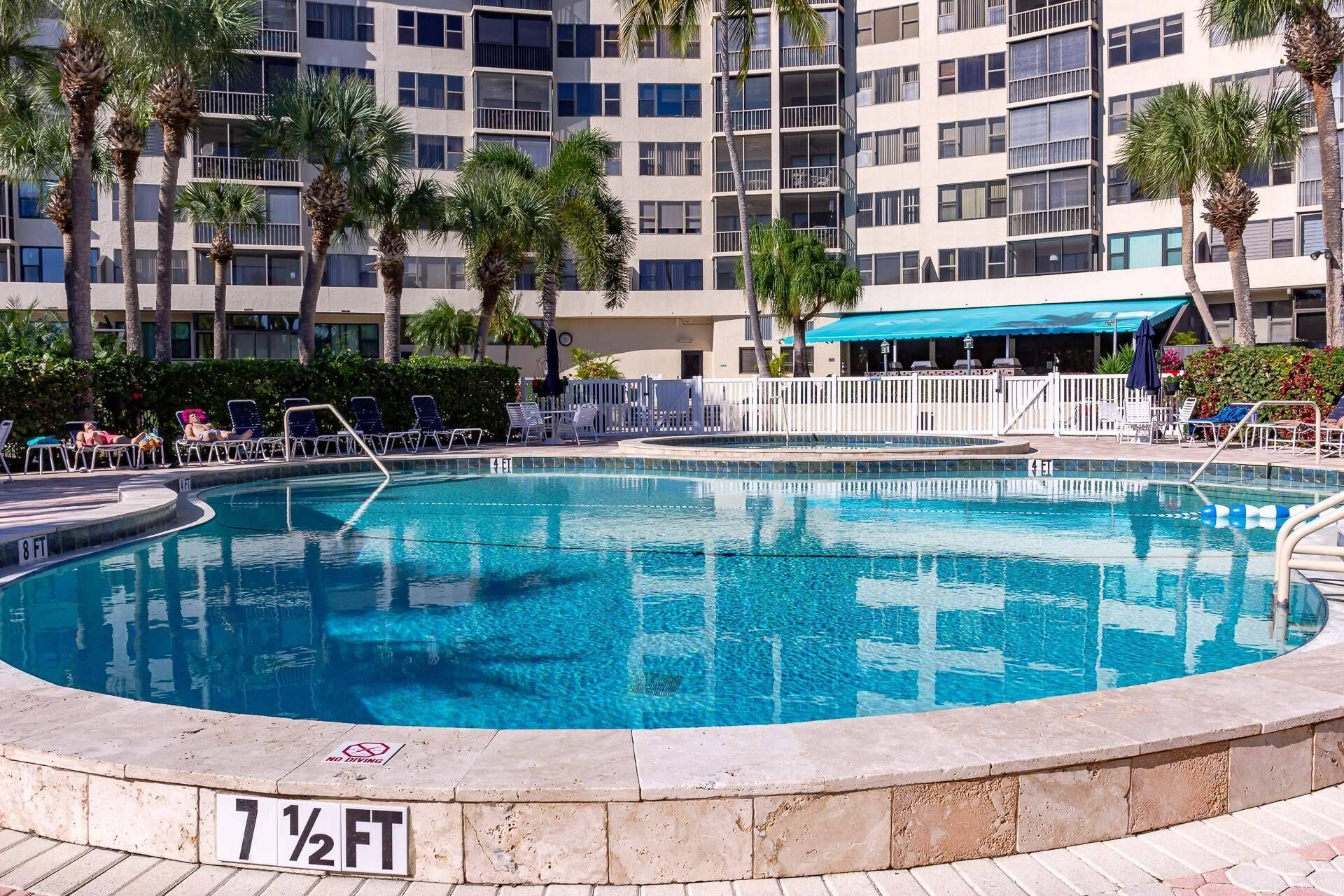 A swimming pool with a no diving sign, surrounded by lounge chairs and palm trees, with a multi-story building in the background.