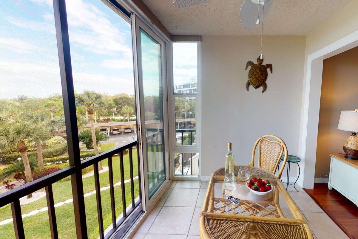A balcony with a view of a landscaped area, a wicker table with a bottle and glass, and a chair.