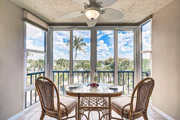 A room with a glass table and chairs, a ceiling fan, and large windows with a view of palm trees and a clear sky. - Resort Style, Heated Pool, Walk to Top Beach (Siesta Key)