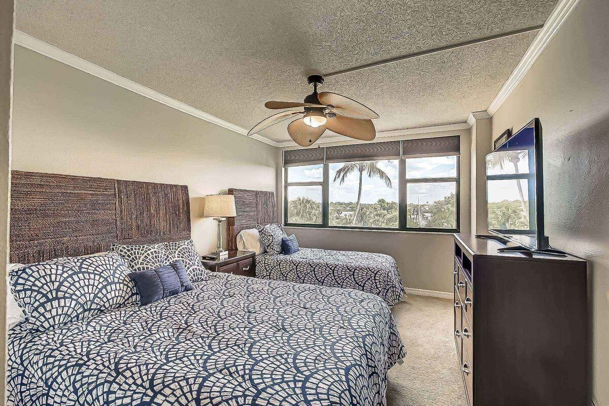 A hotel room with two beds, a ceiling fan, a window with a view, and a TV.
