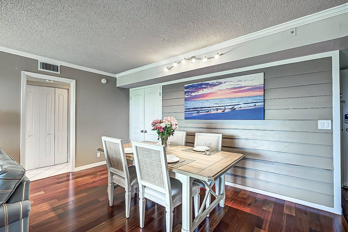 A dining area with a wooden table and white chairs, a painting of a sunset over the ocean on the wall, and a door to the left.