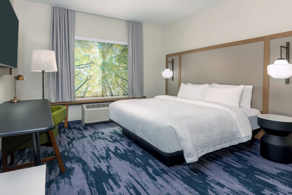 A hotel room with a bed, a desk, a chair, a window with a view of trees, and a wall-mounted lamp.