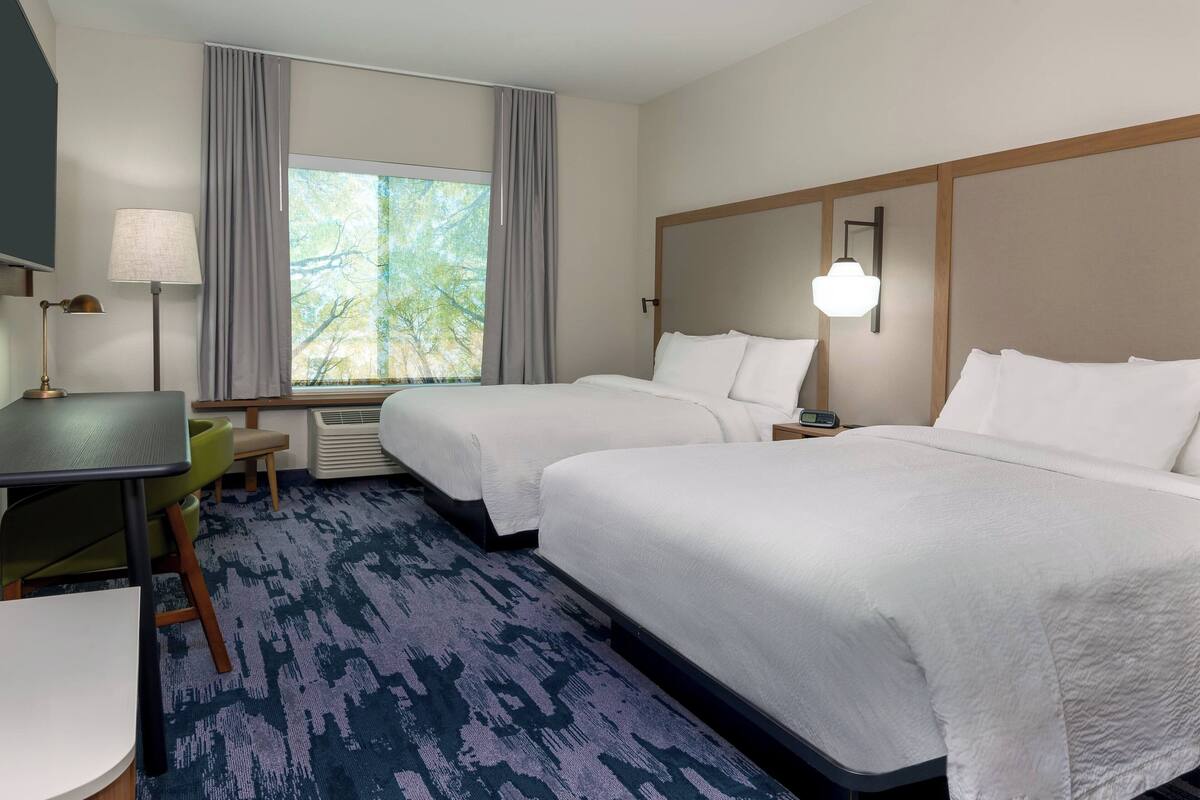 A hotel room with two beds, a desk, a chair, a lamp, and a window with curtains.
