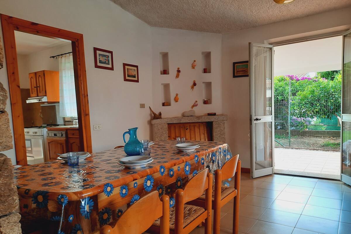 Comedor con mesa lista para cuatro, cocina al fondo y puerta corrediza de vidrio que da a un jardín.