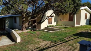 A small, single-story building with a sloped roof, a large tree in front, and a well-maintained lawn. - Villette Sirenella (Aglientu)