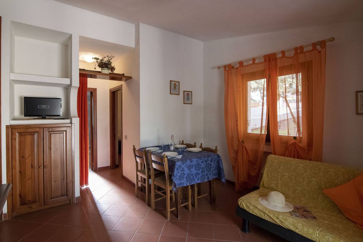 Un comedor con una mesa lista para cuatro personas, un televisor sobre un mueble bajo y un sofá con un sombrero.
