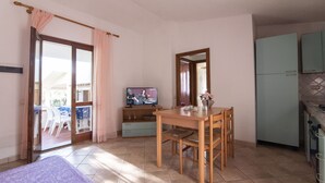 A room with a bed, a dining table with chairs, a television, and a kitchen area. - Villette Sirenella (Aglientu)