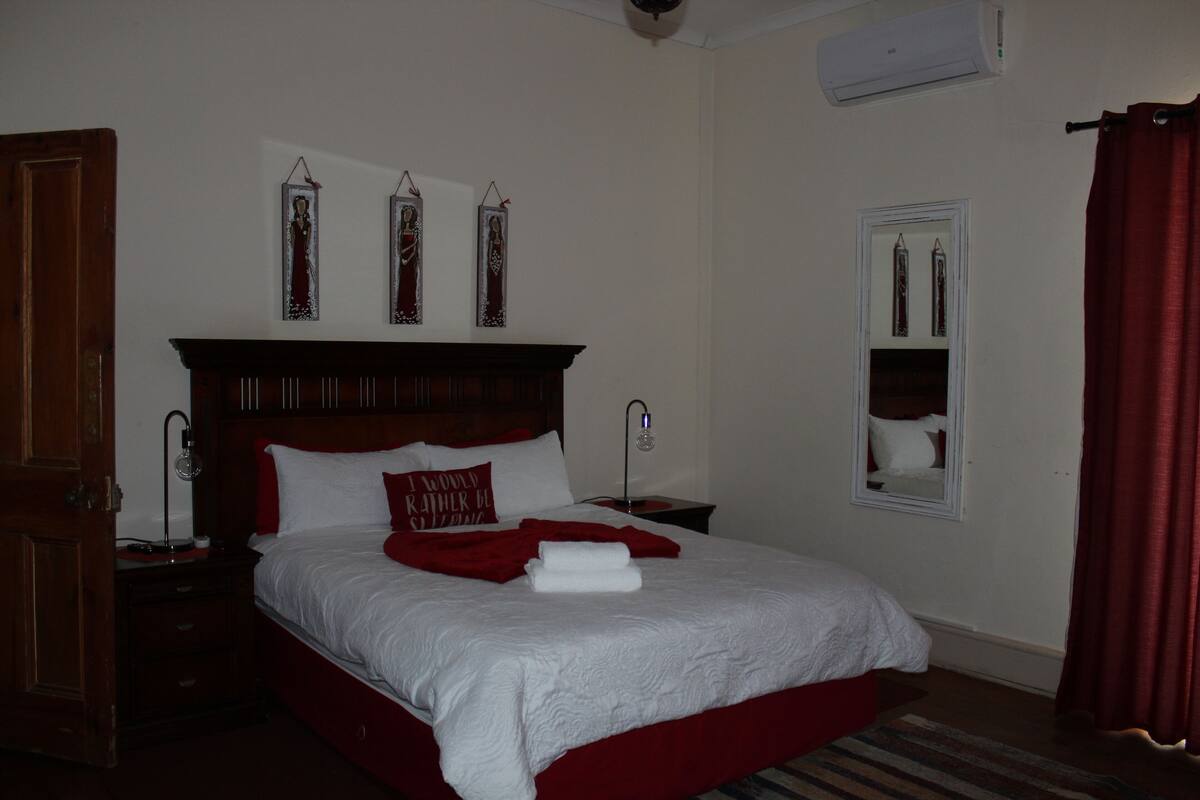 Uma cama bem arrumada com roupa de cama branca e vermelha, uma cabeceira de madeira e uma pequena mesa de cabeceira com uma luminária.