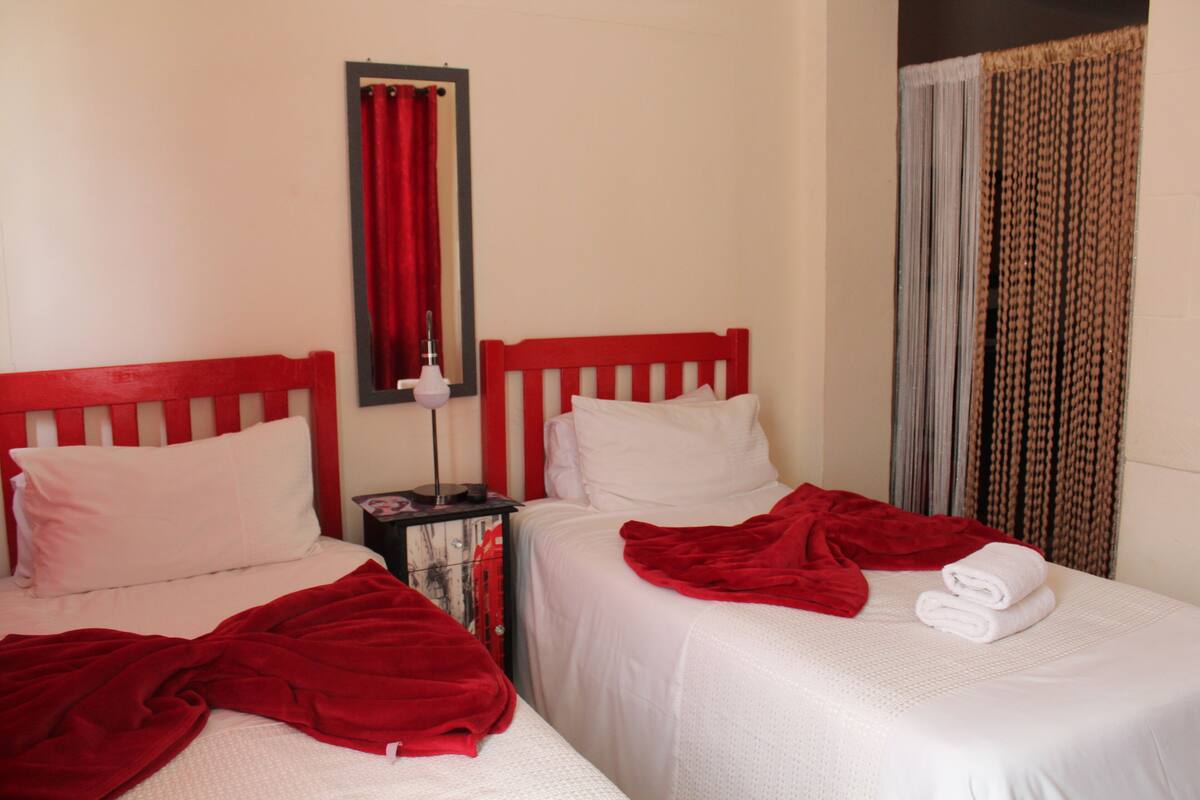 Quarto de hotel com duas camas, cada uma com cabeceira vermelha, roupa de cama branca e um cobertor vermelho em uma das camas. Há uma mesa de cabeceira com uma luminária e uma pequena mesa com um objeto decorativo.