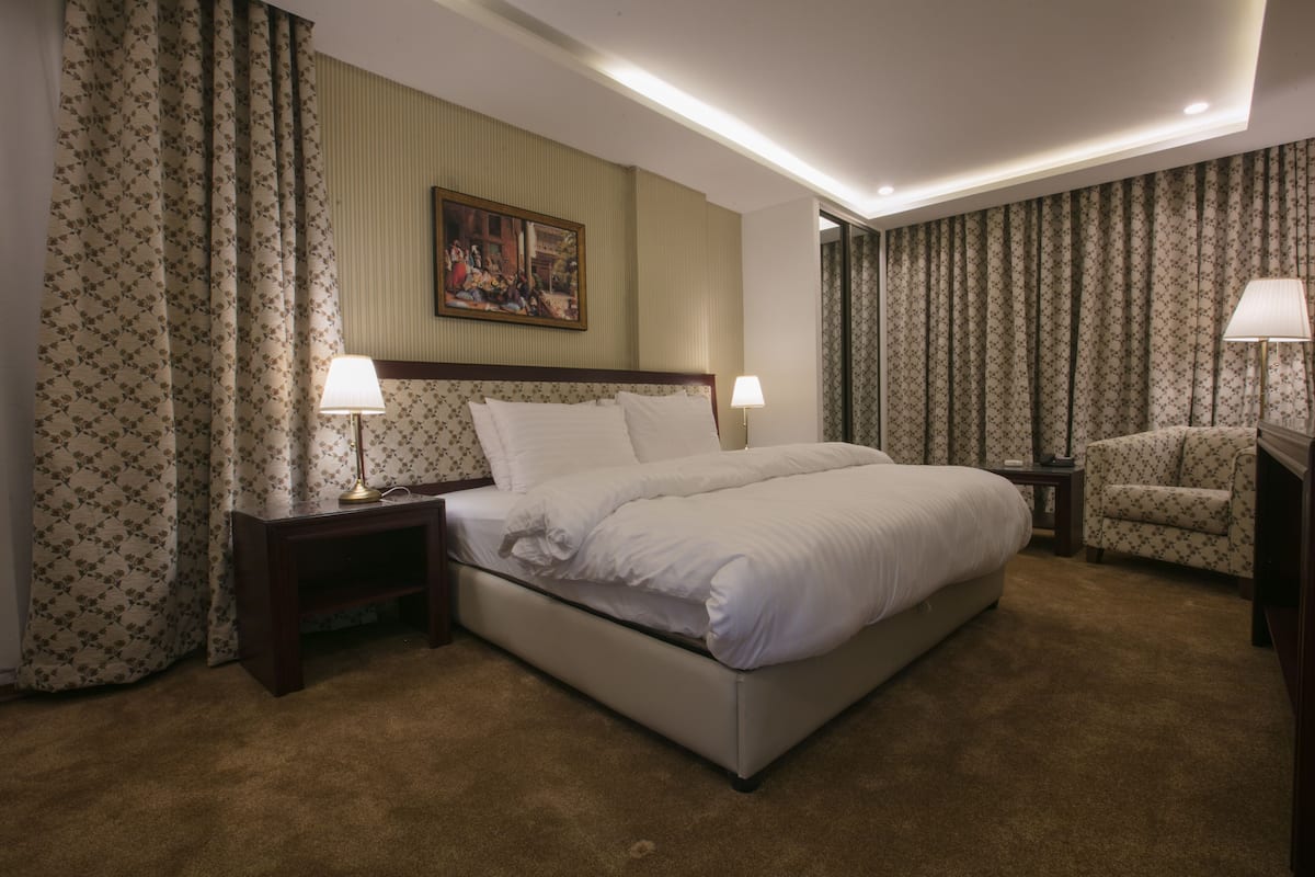 A hotel room with a large bed, two bedside tables with lamps, a chair, and floral curtains.