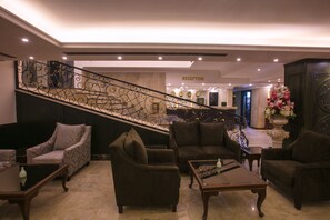 A lobby with a reception desk, a staircase with ornate railings, and several seating areas with sofas and armchairs.