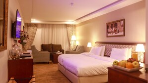 Une chambre d’hôtel avec un grand lit, deux fauteuils, un bureau avec une lampe, une télévision et un tableau au mur.