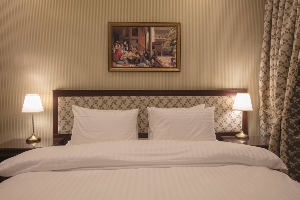 A hotel room with a large bed, two bedside lamps, a framed painting above the bed, and floral-patterned curtains.