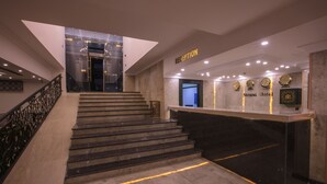 Un espace d'accueil avec un escalier en marbre menant à un comptoir de réception, une enseigne indiquant « RÉCEPTION » et une enseigne « Hôtel Strand » au-dessus du comptoir.