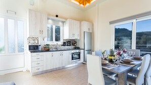 A modern kitchen with light wood cabinets, a central island, and a dining area with a large window. - Creta  Galini  House (Chania Creta)