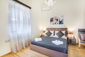 A neatly made bed with a grey duvet, two bedside tables with lamps, a chair, and artwork on the wall.