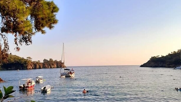 Paisaje costero con barcos en aguas tranquilas, una playa con sombrillas y un cielo despejado.