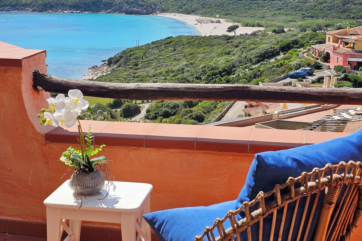 A balcony with a view of a beach and a village. There is a wicker chair and a small table with a flower vase.