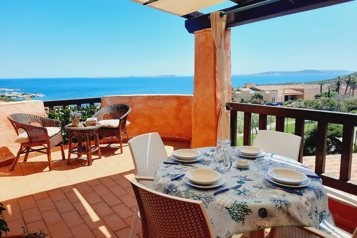 A terrace with wicker chairs and a table set for dining, overlooking a coastal view.