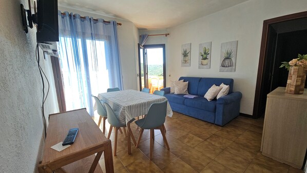 Una sala de estar con un sofá azul, una mesa de comedor con sillas y un televisor montado en la pared.