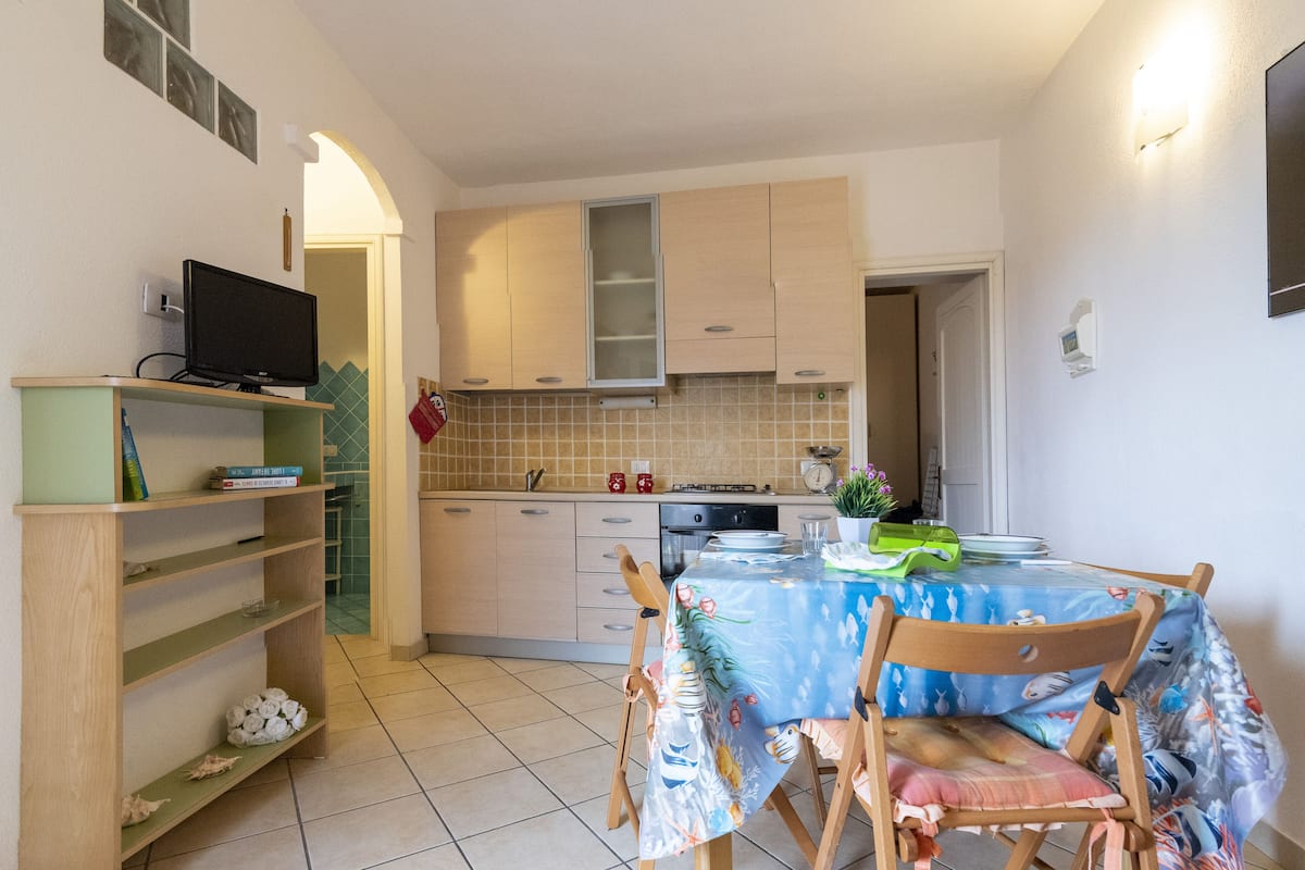 A compact kitchen with a dining area, featuring a table with chairs, a shelf with books, and a television on a stand.