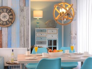 Un coin repas avec une table en bois, des chaises bleues, un panier de fruits et une horloge décorative au mur.