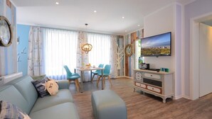 En stue med sofa, skærmpude og spisebord med stole. Der er et tv ophængt på væggen og et lille skab med pynteting.