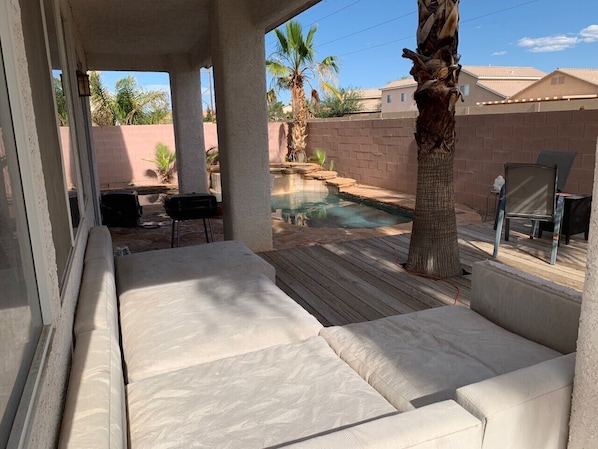 A covered outdoor seating area with a pool, palm trees, and a brick wall in the background.