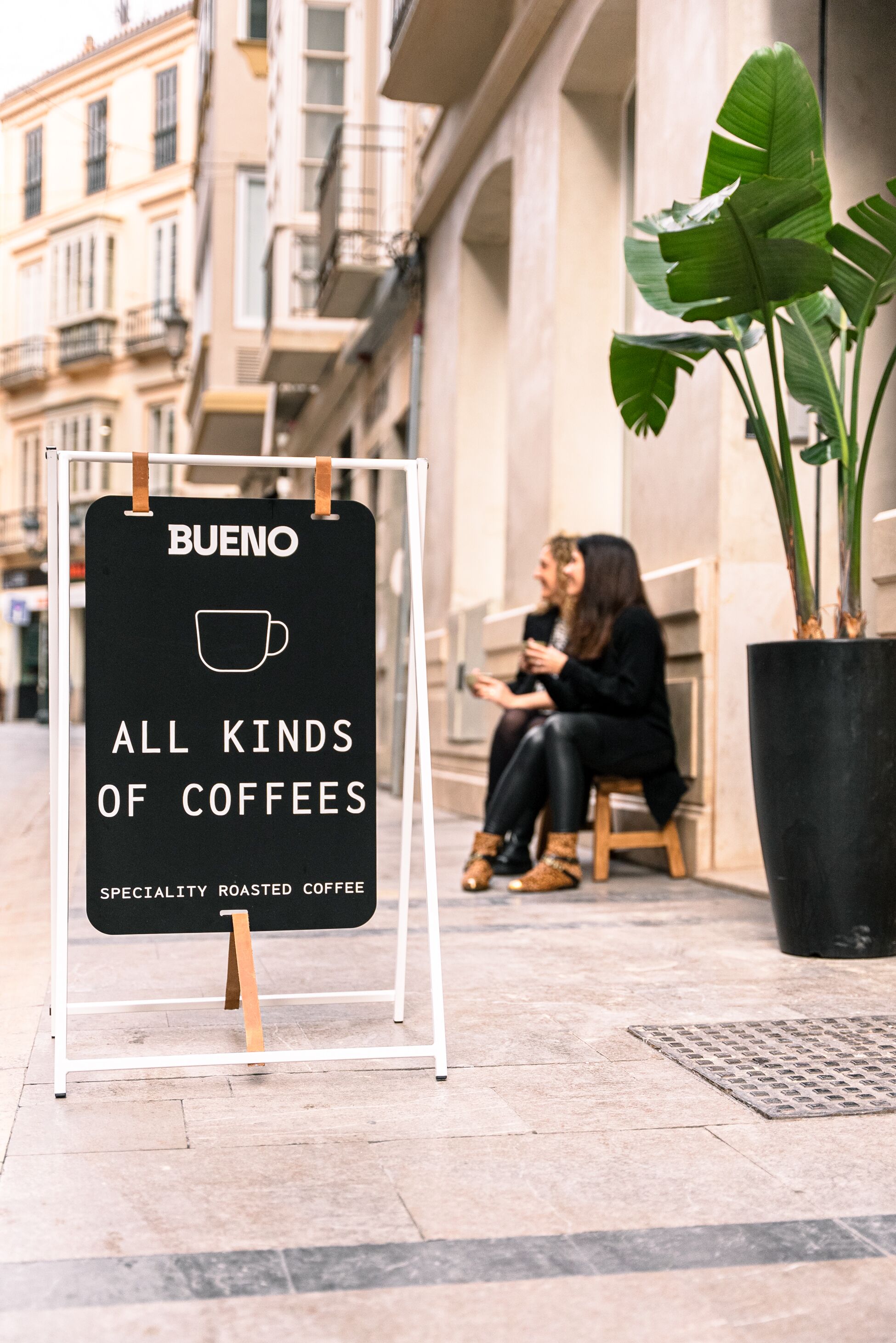 Un letrero anuncia 'BUENO ALL KINDS OF COFFEES SPECIALITY ROASTED COFFEE'. Se ve una pareja sentada en un banco al fondo.
