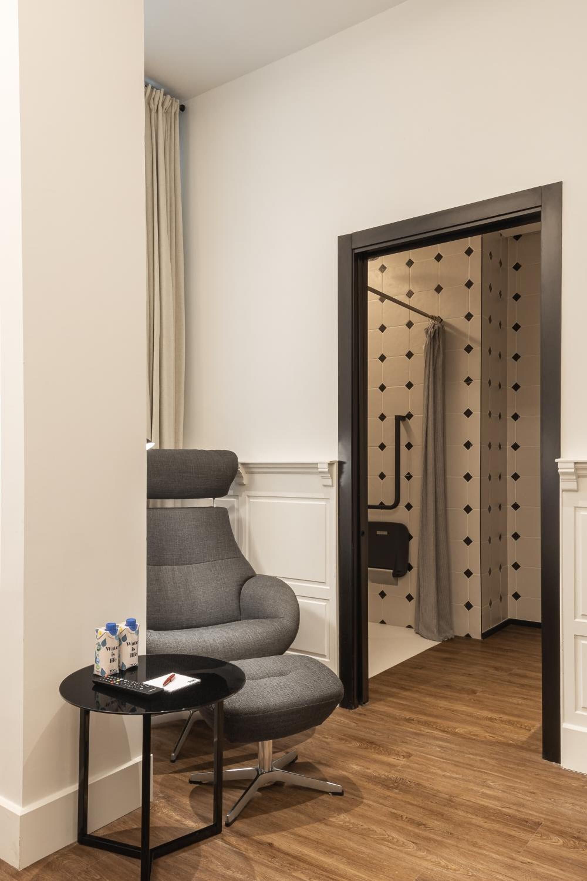A modern room with a grey armchair, a small black table with a remote control, and a white door leading to a bathroom with patterned tiles.