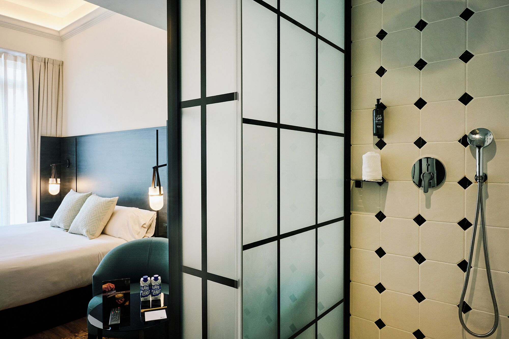 Habitación de hotel con cama, escritorio con computadora portátil, silla, mesita de noche con lámpara y zona de ducha con toalla y dispensador de jabón.