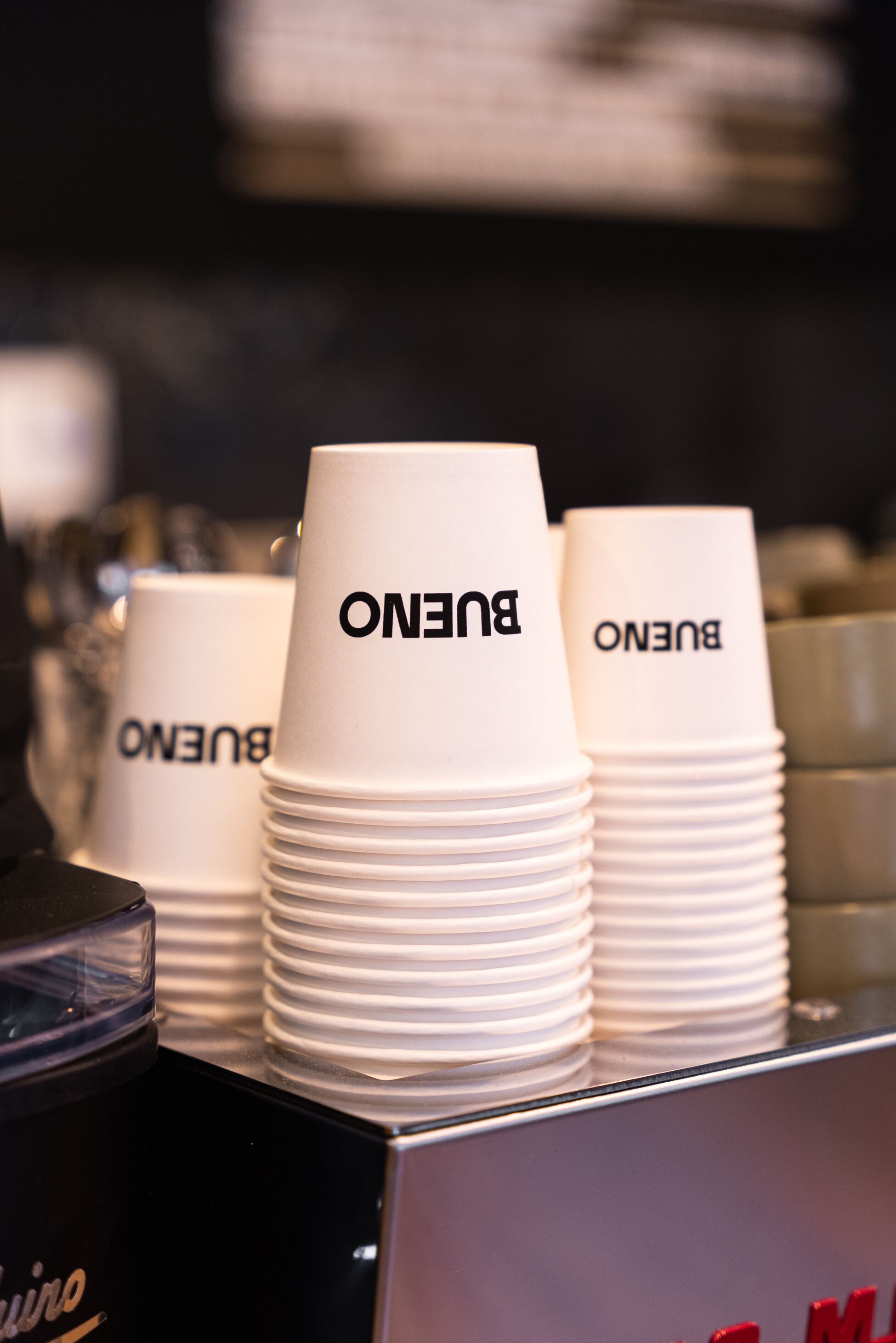 Stacks of white paper cups with "BUENO" printed on them, arranged on a counter.