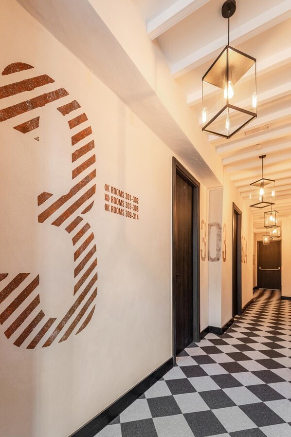 Un couloir d’hôtel avec un sol à carreaux, un grand chiffre « 3 » peint sur le mur et un luminaire aux formes géométriques.