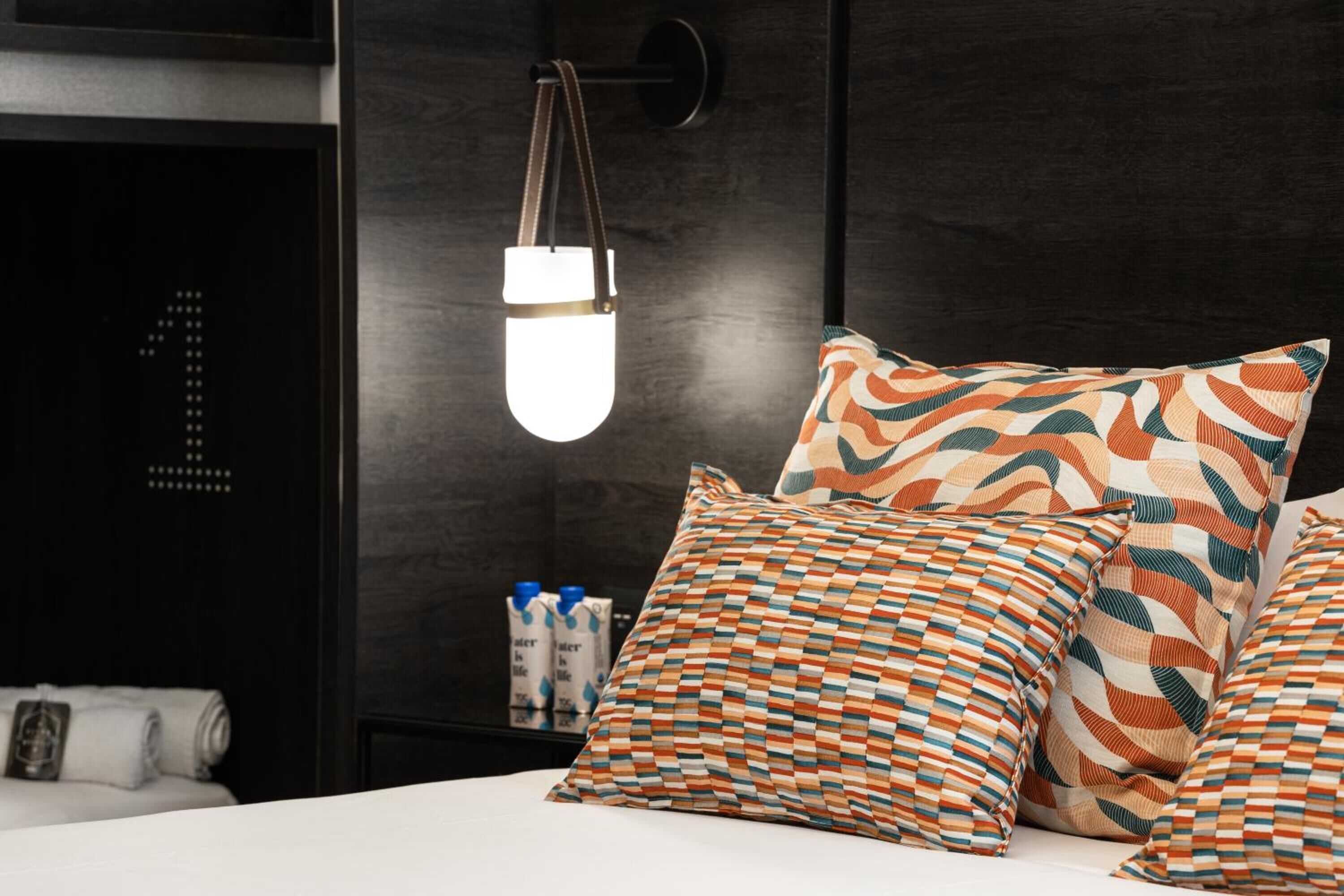 Habitació d'hotel amb un llit, almoinades estampades, una taula de nit amb ampolles i un llum penjada a la paret.
